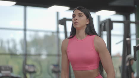 A-brunette-woman-in-a-pink-suit-alternately-raises-dumbbells-to-the-top-in-front-of-her-while-training-her-shoulders-in-the-gym.-Standing-exercise-for-training-the-shoulders-and-arms.-Weight-training